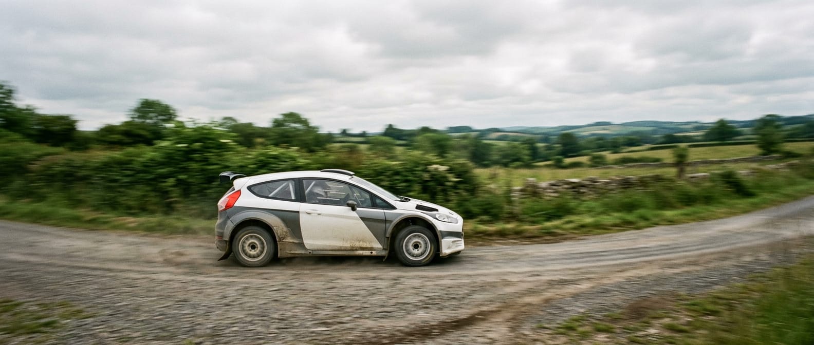 A white rally car panning past hedgerows on a gravel road